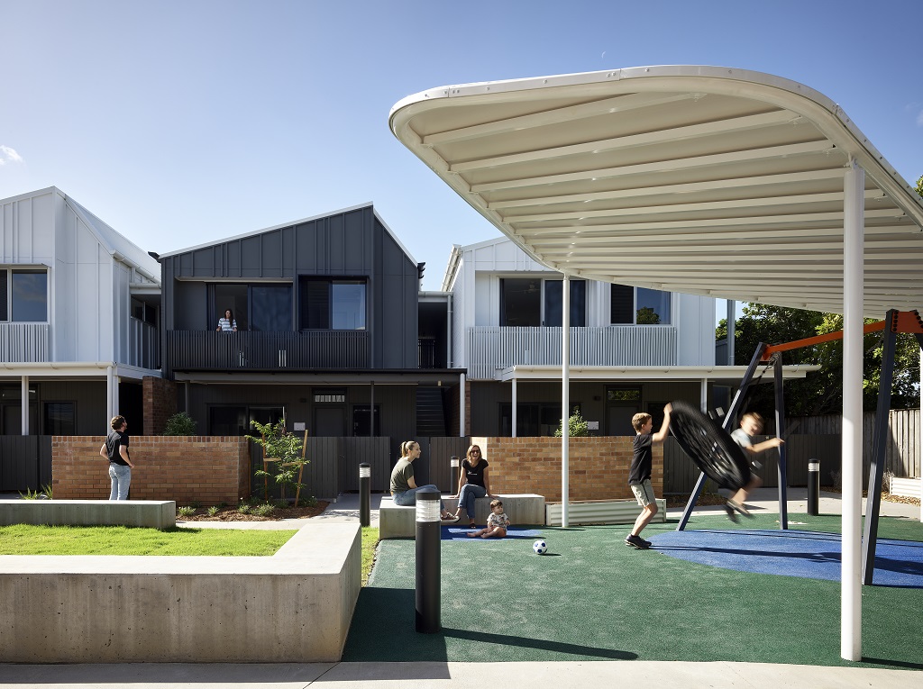 View of the generous communal space, showing concrete bench seats, covered area, and a swing. View of the townhouse-style apartments behind. 